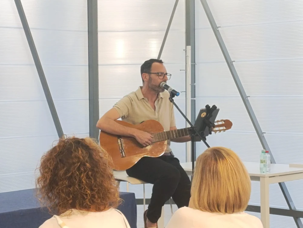 Interpretación con guitarra ante el público, junto a unos ventanales.